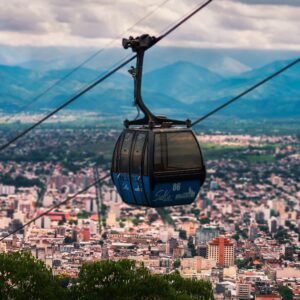 Ropeway moving over green trees growing near city buildings located under sky with clouds